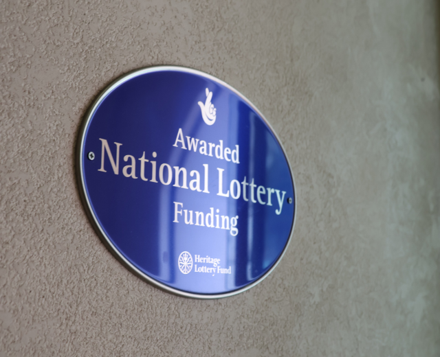 Blauer Schild an einer Gebäudewand mit der Aufschrift "National Lottery Funding" in fetter schwarzer Schrift auf weißem Hintergrund.