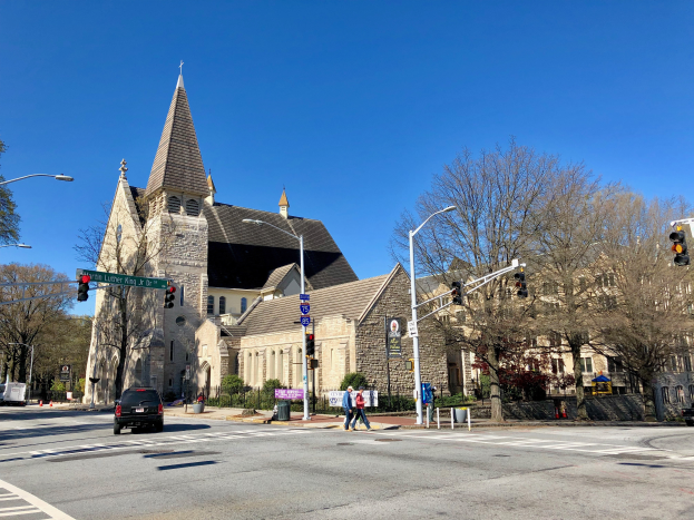 St. Luke's Episcopal Church, eine große Kirche mit einem Kirchturm, steht an einer Straßenecke umgeben von Gebäuden, Verkehrszeichen, Fahrzeugen, Fußgängern und Grün unter einem klaren blauen Himmel.