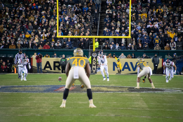 Ein Fussballspiel wird in einem Stadion mit Spielern in Sportkleidung und Helmen im Vordergrund und einer Zuschauermenge im Hintergrund sowie sichtbaren Pfosten und Tafeln mit Text gespielt.