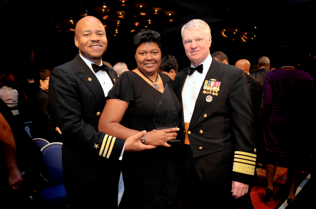 Zwei Männer und eine Frau stehen auf einem roten Teppich; die Frau hält einen Preis, alle drei sind formell gekleidet, mit anderen und beleuchteter Decke im Hintergrund.