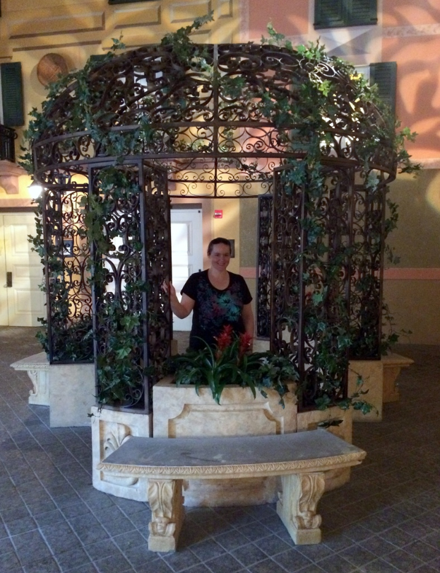 Eine Bank im Vordergrund mit einer verzierten Eisen Säule und Kletterpflanzen dahinter, eine Frau in einem schwarzen Kleid, die in der Säule steht, und ein Gebäude mit Wänden, Fenstern und Türen im Hintergrund.