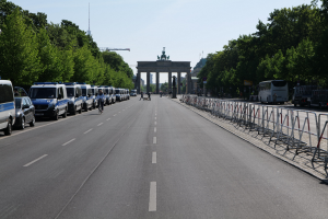 Lange Reihe von Polizeiwagen vor dem Brandenburger Tor geparkt, mit Radfahrern und Passanten, Absperrungen, Bäumen, ein Tor mit Statuen im Hintergrund und wolkenloser Himmel.