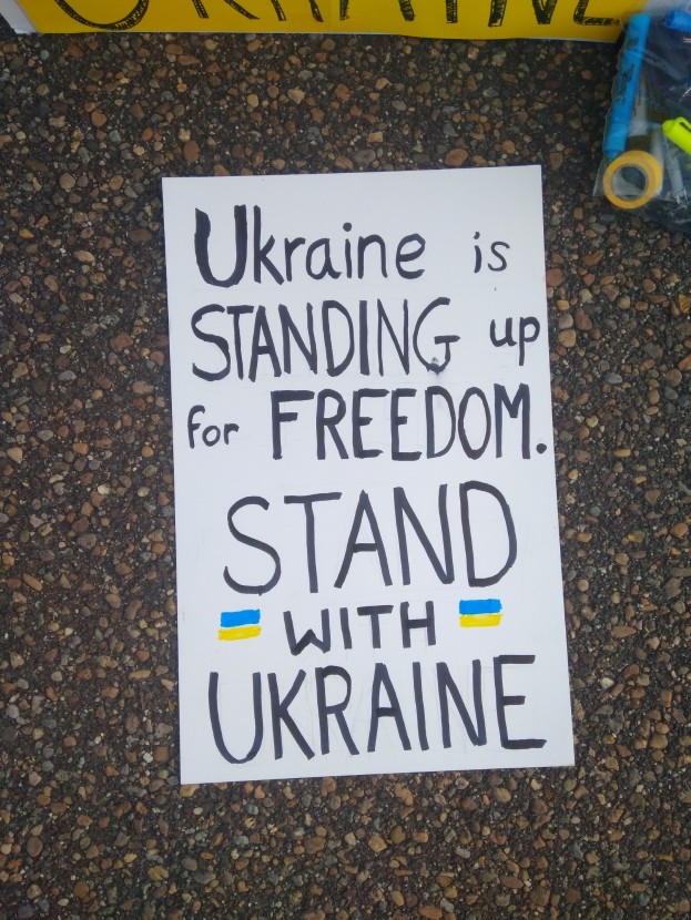 Weißer Zettel mit dem Text "Ukraine steht für Freiheit, steht mit Ukraine" auf dem Boden vor einer gelben Fahne, mit einer Plastikhülle rechts mit verschiedenen Gegenständen.