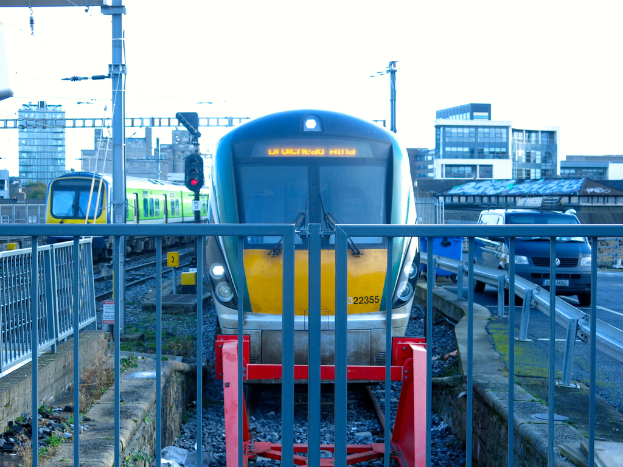 Zwei Züge auf der Schiene mit einem Zaun im Vordergrund, Pfosten auf dem Land, einer mit einer Signalleuchte, ein Auto auf der Straße rechts, Gebäude im Hintergrund und der Himmel oben.