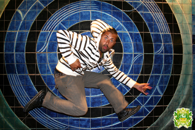 Ein Mann tanzt vor einem bunten Hintergrund und einem Logo mit Text in der rechten oberen Ecke.