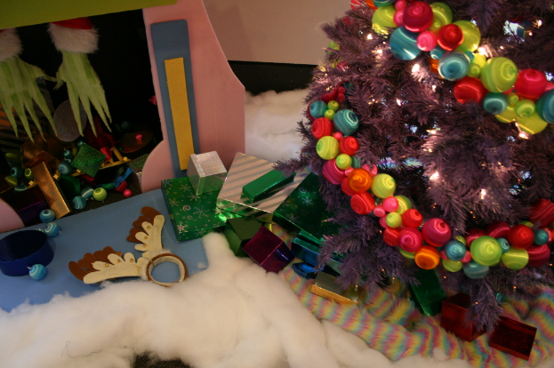 Eine Indoor-Szene mit einem geschmückten Weihnachtsbaum rechts, Geschenkboxen und Dekorationsgegenständen in der Mitte und einer kleinen Hütte mit Dekorationen oben links, mit Watte und Wolle am Boden.