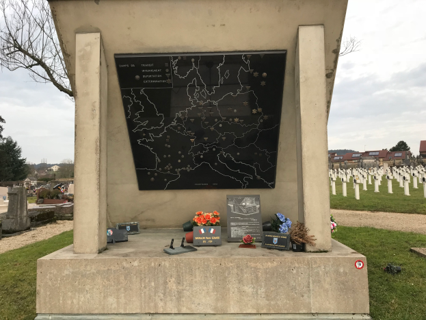 Ein Denkmal auf einem Friedhof mit einer Karte darauf, umgeben von Blumensträußen und anderen Gegenständen, mit Gräbern, Bäumen, Gebäuden und einem klaren blauen Himmel im Hintergrund, das Grab eines deutschen Soldaten, der im Zweiten Weltkrieg getötet wurde.