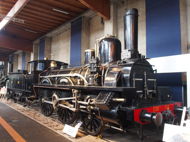 Alte Dampflokomotive in einem Museums Setting mit umgebenden Eisenbahnschienen, Informationsschildern und anderen Ausstellungen, vor einer Wand mit Deckenleuchten.