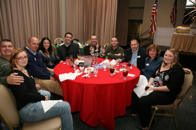 Eine Gruppe strahlender Menschen, die an einem Tisch mit Gläsern, Papieren, Weinflaschen und Flagge sitzen.
