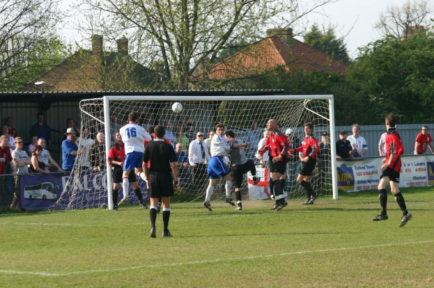 Spieler spielen Fußball auf einem Feld mit einem Tor, während Zuschauer dahinter stehen; Bäume und Häuser sind im Hintergrund zu sehen.
