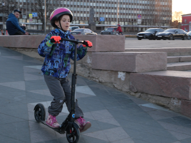 Ein junger Junge in Helm und Handschuhen fährt mit einem Roller eine Treppe hinunter, vorbei an Fahrzeugen, Menschen, Bäumen, Pfählen, Brettern, Gebäuden und einem klaren blauen Himmel.