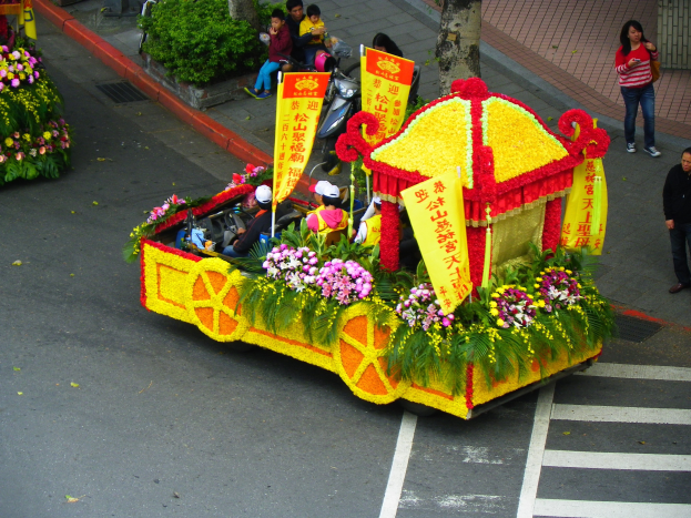 Ein Paradewagen mit Blumen und Bannern geschmückt trägt Menschen auf seinem Dach, während Zuschauer rechts daneben stehen und Bäume im Hintergrund zu sehen sind.