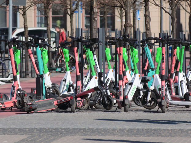 Reihe von Elektro-Rollern auf einer Straße geparkt mit einer Person im Vordergrund, unscharfer Hintergrund von Bäumen, Masten, Plakaten, Fahrzeugen und Gebäuden.