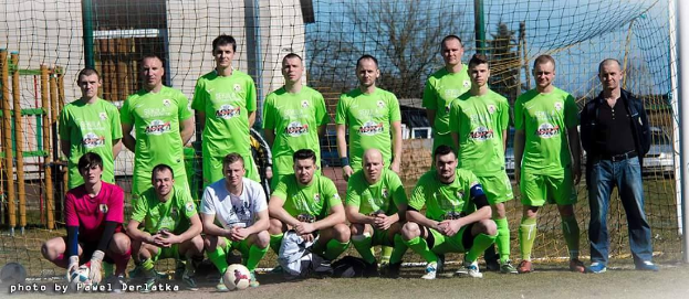Eine Gruppe von Männern in grünen T-Shirts steht und hockt auf einem Fußballfeld mit einem Ball vor ihnen, ein Torpfosten rechts und ein Hintergrund aus Bäumen, Gebäuden, Mästen, Fahrzeugen und dem Himmel, mit Text unten links, der die Nummern von 1 bis 72 auflistet.