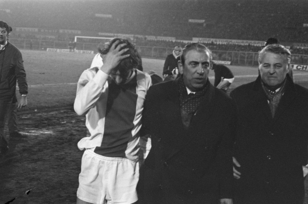 Eine Gruppe von Männern steht zusammen auf einem Fußballfeld, sieht frustriert aus, mit einem Stadion im Hintergrund, in Schwarz-Weiß aufgenommen.