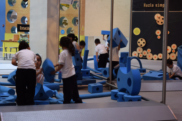 Kinder beim Spielen in einer Turnhalle mit Gegenständen in den Händen und einer Texttafel an der Wand sowie Stangen rechts daneben.