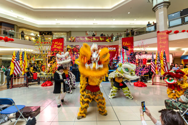 Lebendiges chinesisches Neujahrsfest in einem Einkaufszentrum mit tanzenden Menschen in Kostümen, anderen auf Stühlen sitzend und mobile Telefone haltend sowie verschiedenen Dekorationen, Flaggen und Bannern im Hintergrund.