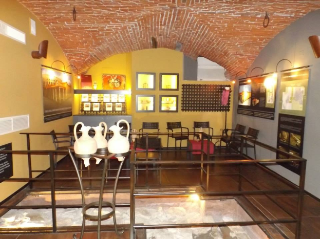 Ein geräumiger Museumsraum mit mehreren Tischen, Stühlen, Displaytafeln, gerahmten Kunstwerken an den Wänden, Vasen, Leuchten und anderen Ausstellungsstücken.