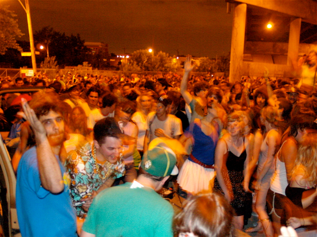 Menschen tanzen vorne bei einem Konzert mit Lichtern, Masten, B├Ąumen und anderen Gegenst├Ąnden im Hintergrund.