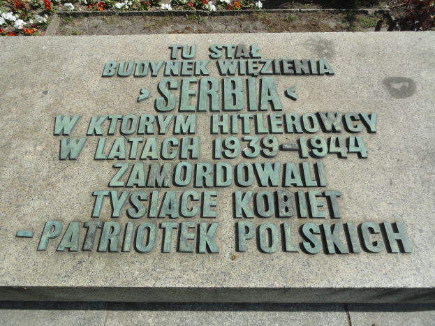 Denkmalstein mit einer Tafel vor einem Gebäude umgeben von blühenden Pflanzen.