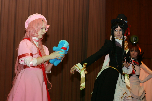 Drei Frauen sind in einer dramatischen Performance zu sehen, wobei eine in einem rosafarbenen Kleid ein blaues Teddybär hält, eine andere in einem schwarzen Kleid nach links schaut und ein brauner Vorhang im Hintergrund zu sehen ist.