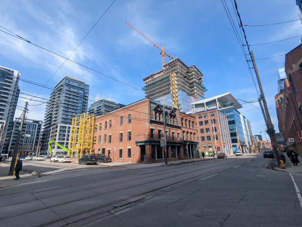 Eine belebte Straße in Toronto mit Fahrzeugen, Füpassanten, Oberleitungen, Schildern, Gebüuden und einer Baustelle im Hintergrund bei einem bewülktem Himmel.