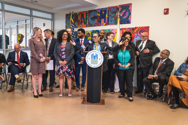 Gruppe von Menschen vor einem Rednerpult mit einem Mikrofon und einer Tafel mit der Aufschrift "Sancho City Councilman Joins City Council", einige sitzen auf Stühlen und andere stehen, mit Flaggen, einer Tafel, einem Feuerlöscher und Deckenleuchten an der Rückwand.