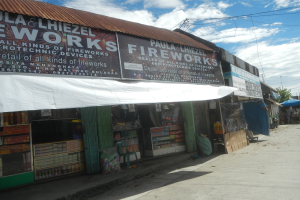 Ein Ladengeschäft mit einer "Feuerwerk"-Schilder, umgeben von verschiedenen Gegenständen und Gebäuden, mit Bäumen, Masten, Drähten und einem bewölktem Himmel im Hintergrund.