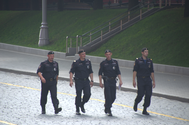 Vier Polizisten in Uniform gehen eine Straße entlang und halten Papiere in der Hand, mit einem Gebäude, Bäumen und einem Pfahl im Hintergrund.