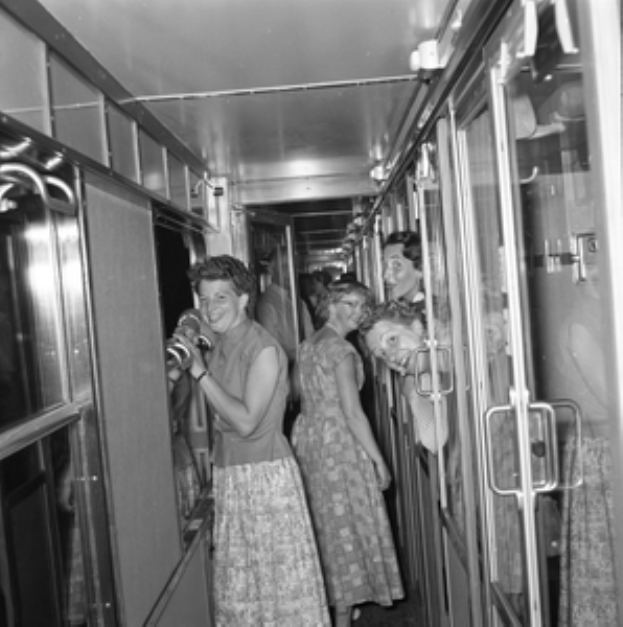 Eine Gruppe von Frauen, die auf einem Zug mit Türen auf beiden Seiten und Lichtern öber ihnen steht, in Schwarzweiß dargestellt.