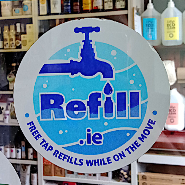 Ladenfront mit einem "Refill ie - Kostenlose Zapfrefills unterwegs"-Schild im Vordergrund und Regalen mit Flaschen und Artikeln im Hintergrund.