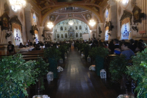 Eine Hochzeitszeremonie in einer Kirche mit Gästen auf Bänken sitzend und im Gang stehend, dekoriert mit Zimmerpflanzen und Schmuckstücken, sowie Vorhängen an den Fenstern und hängenden Kronleuchtern.