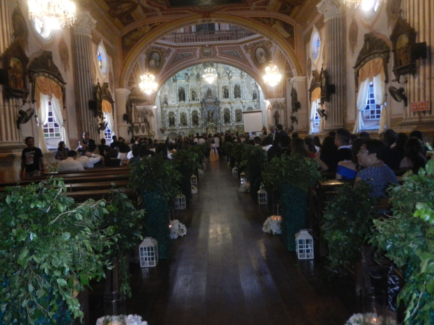 Eine Hochzeitszeremonie in einer Kirche mit Gästen auf Bänken sitzend und im Gang stehend, dekoriert mit Zimmerpflanzen und Schmuckstücken, sowie Vorhängen an den Fenstern und hängenden Kronleuchtern.