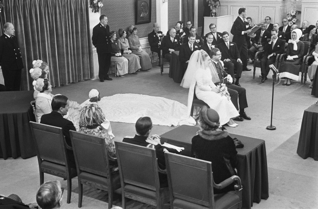 Schwarzes und weißes Foto einer Hochzeitszeremonie mit Menschen, die sitzen und stehen, einem Mann mit Mikrofon, einer Frau mit Blumenstrauß, einem Podium mit Mikrofon auf der rechten Seite, Blumengestecken und einem Vorhang auf der linken Seite, einem gerahmten Foto an der Wand und einer Tür im Hintergrund.