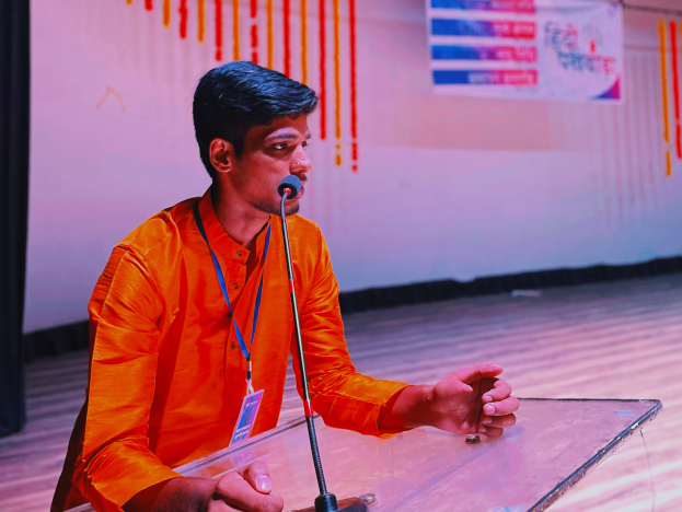 Ein Mann in einem orangen Hemd und mit Ausweis sitzt an einem Rednerpult mit Mikrofon und spricht vor einem weißen Bildschirm mit Banner und schwarzem Vorhang links.