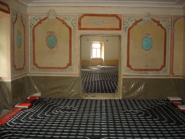 Ein Raum mit einer Fußbodenheizung in der Mitte, umgeben von Wänden und einem Fenster, mit verschiedenen Gegenständen auf dem Boden, die mit dem Heizungssystem in Zusammenhang stehen.