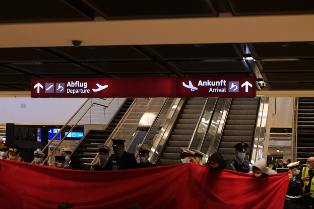Eine Gruppe von Menschen mit M├╝tzen und Masken steht vor einer Rolltreppe am Flughafen, mit einem roten Tuch in der Hand und positioniert unter Schildern, wahrscheinlich beim Einsteigen ins Flugzeug w├Ąhrend der Coronavirus-Pandemie.