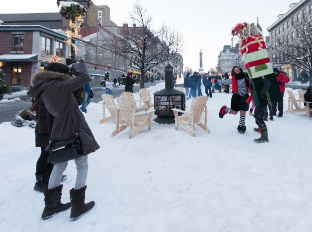 Eine Gruppe von Menschen steht auf Schnee mit Stühlen und Bäumen im Vordergrund, Gebäude im Hintergrund und der Himmel darüber.