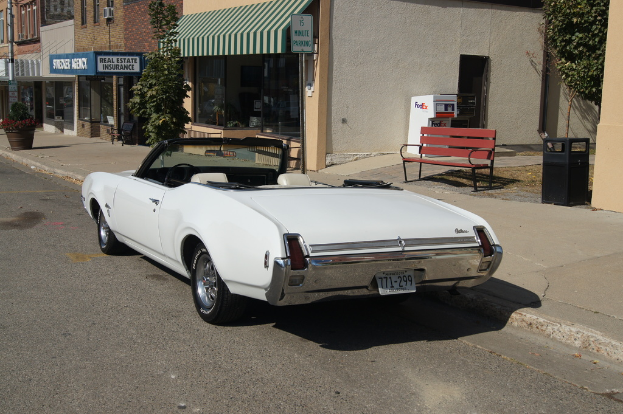 Ein weißer Wagen mit dem Kennzeichen 771-299 steht auf der Straße, daneben eine Bank, im Hintergrund ein Gebäude und ein Geschäft namens 'Immobilienversicherung'.