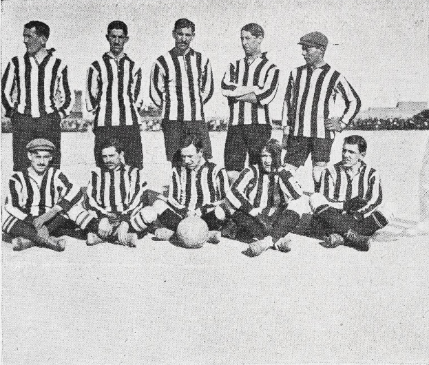 Ein Schwarz-Weiß-Foto einer Fußballmannschaft, einige stehen und einige sitzen auf dem Boden mit einem Ball davor, mit Gebäuden und einem klaren Himmel im Hintergrund und Text unten.