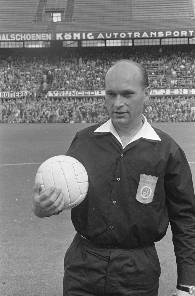 Ein Schwarz-Weiß-Foto eines Schiedsrichters auf einem Fußballfeld, der einen Ball hält, mit Zuschauern und Tafeln mit Text im Hintergrund.