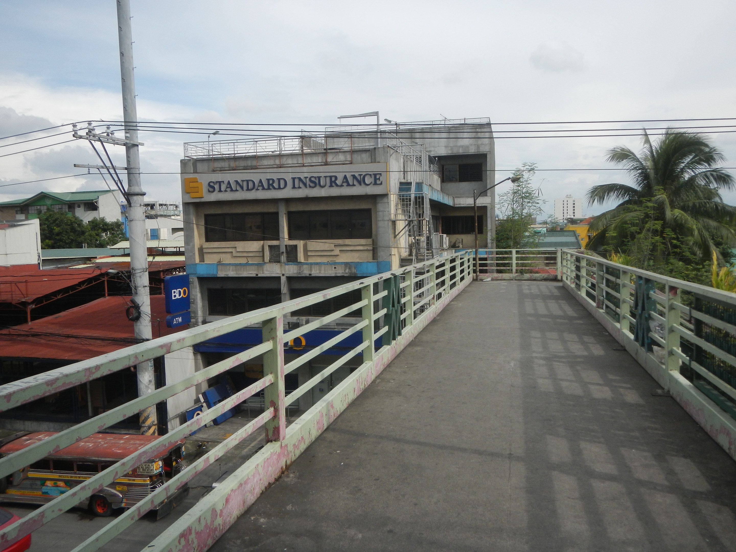Eine Brücke mit Geländern, Fahrzeugen, Gebäuden, Bäumen, Strommasten mit Drähten und einem bewölkten Himmel im Hintergrund, mit einem Gebäude an der Seite mit einer "Standard Insurance"-Aufschrift.