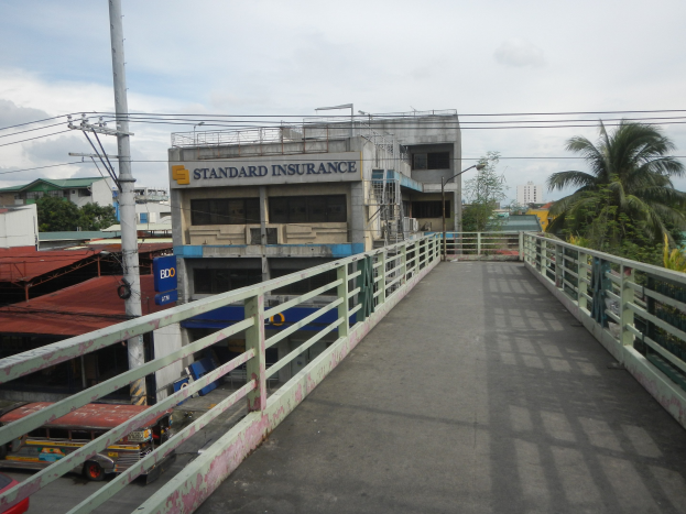 Eine Brücke mit Geländern, Fahrzeugen, Gebäuden, Bäumen, Strommasten mit Drähten und einem bewölkten Himmel im Hintergrund, mit einem Gebäude an der Seite mit einer "Standard Insurance"-Aufschrift.