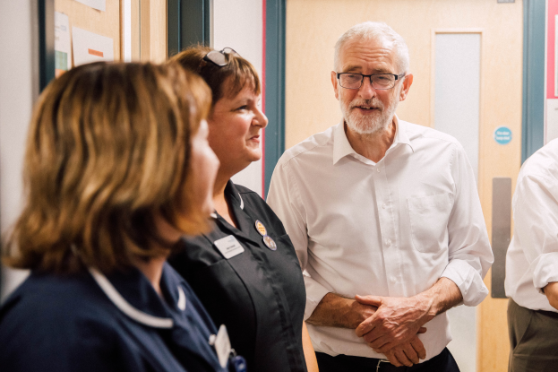 Eine Gruppe von Menschen, die in einem Raum mit einer Tür im Hintergrund und einer Anschlagtafel mit Papieren an der linken Seite steht und spricht.