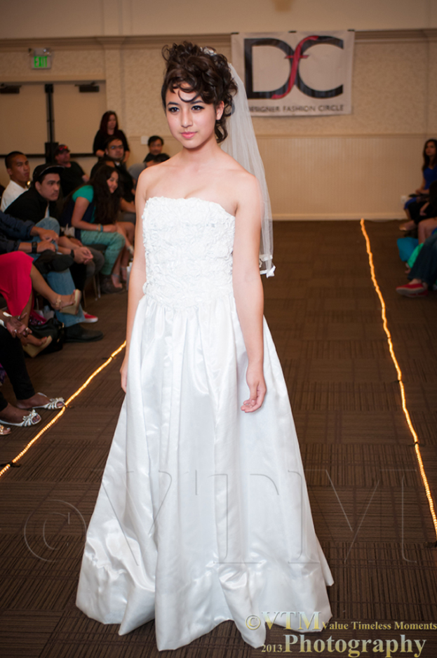 Eine Frau in einem weißen Kleid lächelt und posiert vor einem weißen Banner mit Text, während Menschen auf Stühlen auf beiden Seiten von ihr sitzen und ein brauner Boden unten sichtbar ist.