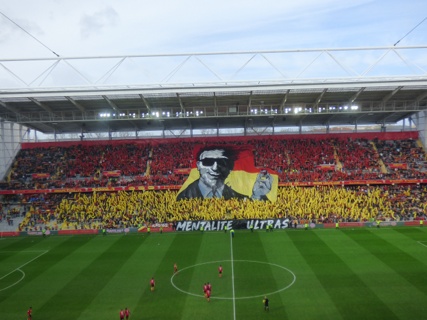Ein Fußballstadion voller Menschen auf dem Feld und auf den Tribünen, mit einem zentralen Banner mit einem Porträt und Text, unter einem sichtbaren Himmel, Heimstadion von Watford Football Club.