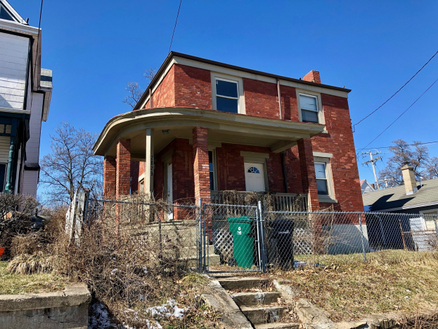 Ein Ziegelhaus mit einer vorderen Einfriedung, umgeben von Gras, Pflanzen, Bäumen und Mülltonnen, mit einer Treppe, die hinaufführt, und Strommasten und -leitungen im Hintergrund.