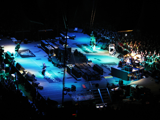 Menschen, die auf einem Podium mit Kästen, Lautsprechern, Lichtern und Mikrofonen performen, mit einer großen Menge auf beiden Seiten und einem dunklen Hintergrund.