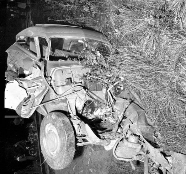Ein Schwarz-Weiß-Foto eines schwer beschädigten Autos am Straßenrand, mit einer zerbeulten Front, zersplitterten Fenstern, einem Baum im Hintergrund und verstreuten Gegenständen um die Unfallstelle.