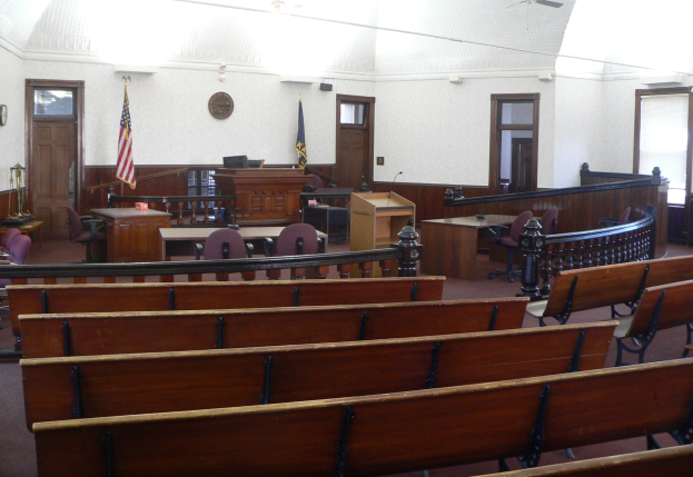 Ein Gerichtssaal mit hölzernen Kirchenbänken, Tischen, Stühlen, einem Podium, Flaggen, Türen, einer Uhr an der Wand und Lampen und Ventilatoren an der Decke.